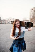 Young teenage urban girl with skateboard, wear on glasses, cap and ripped jeans at the yard sports ground on sunset making selfie on phone. photo