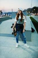 Young teenage urban girl with skateboard, wear on glasses, cap and ripped jeans at skate park on the evening. photo