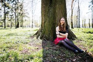 retrato de una chica con maquillaje brillante con labios rojos, collar de gargantilla negra en el cuello y falda de cuero rojo sentada cerca de un árbol en el parque. foto