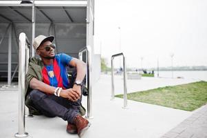 Stylish african american boy wear at cap, football t-shirt and sunglasses sitting near steel railings. Black sports man portrait. photo