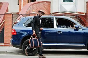 Stylish black man at glasses with hat, wear on suit with handbag against luxury car. Rich african american businessman. photo