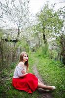 Portrait of sitting beautiful girl with red lips at spring blossom garden on green grass, wear on red dress and white blouse. photo