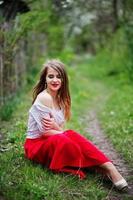 Portrait of sitting beautiful girl with red lips at spring blossom garden on green grass, wear on red dress and white blouse. photo