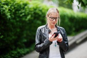 elegante mujer rubia vestida con jeans, gafas y chaqueta de cuero con teléfono a mano, contra arbustos en la calle. retrato de modelo urbano de moda. foto