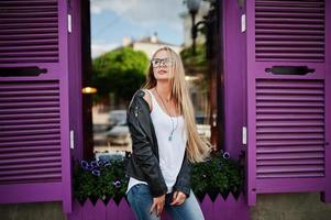 Stylish blonde woman wear at jeans, glasses and jacket posed at street against purple window. Fashion urban model portrait. photo