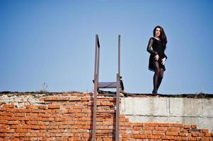 Portrait brunette girl with red lips wearing a black dress posed on the roof near the ladder. Street fashion model. photo