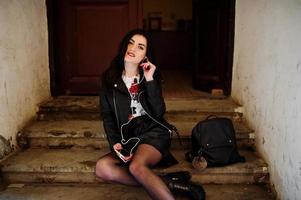 Young goth girl on black leather skirt, jacket and punk shoes sitting on stairs of old house and listening rock music at headphones of mobile phone. photo