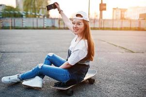 Young teenage urban girl with skateboard, wear on glasses, cap and ripped jeans at the yard sports ground on sunset making selfie on phone.