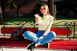 Young teenage girl sitting on bench with ice cream at hands, wear on yellow t-shirt, jeans and sunglasses. photo