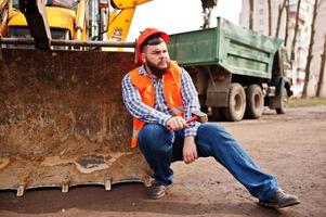 Beard worker man suit construction worker in safety orange helmet, against tractor with hammer at hand. photo