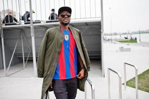 Stylish african american boy wear at cap, football t-shirt and sunglasses posed on steel railings. Black sports man portrait. photo