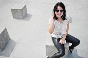 Portrait of brunette girl listening music at headphones from mobile phone, wear on women's leather pants and white blouse, sunglasses, against stone cubes. photo