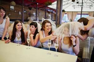 Group of cheerful girls at white shirts sitting at table and drink champagne on hen party. photo