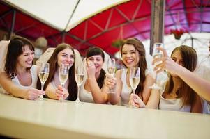 grupo de chicas alegres con camisas blancas sentadas en la mesa y bebiendo champán en la despedida de soltera.