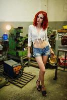 Red haired girl wear on short denim shorts and white blouse with welding mask at hands posed at industrial machine at the factory. photo