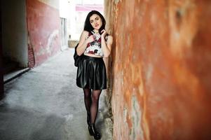 Young goth girl on black leather skirt with backpack posed against grunge wall. photo