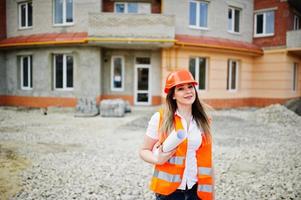 mujer ingeniera constructora con chaleco uniforme y casco protector naranja sostiene papel comercial contra el nuevo edificio. tema de bloque de vivienda de propiedad. foto