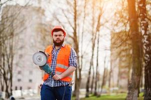 Portrait of brutal beard worker man suit construction worker in safety orange helmet against pavement with angular grinding machine in hand. photo