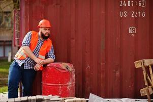 Brutal beard worker man suit construction worker in safety orange helmet near red barrel. photo