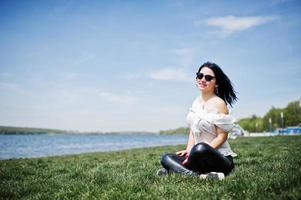 Brunette girl on women's leather pants and white blouse, sunglasses, sitting on geen grass against beach of lake. photo