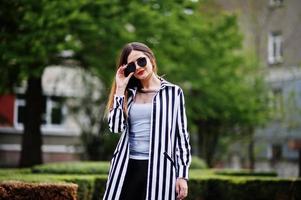 Fashionable woman look with black and white striped suit jacket, leather pants and sunglasses posing against bushes at street. Concept of fashion girl. photo