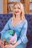 portrait of beautiful attractive cheerful blonde Slavic woman in blue jeans, blue blouse on the blue couch holding globe and dreaming about travelling photo