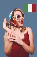 Divertido elegante sonriente alegre elegante mujer rubia con maquillaje y con zapato de tacón en sus brazos y bandera italiana en el fondo foto