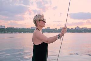 Beautiful blonde happy woman in sunglasses and long black dress enjoying sunset on the yacht in the sea. Travel, adventure, chill, relaxation concept photo