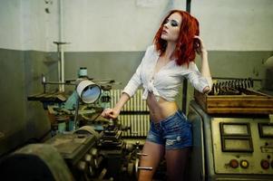 Red haired girl wear on short denim shorts and white blouse posed at industrial machine at the factory. photo