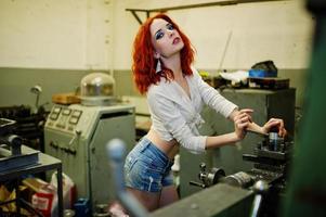 Red haired girl wear on short denim shorts and white blouse posed at industrial machine at the factory. photo