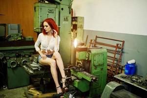Red haired girl wear on short denim shorts and white blouse posed at industrial machine at the factory.