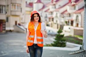 Engineer builder woman in uniform waistcoat and orange protective helmet hold business drawing paper roll against new building. Property living block theme. photo