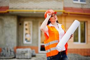 Engineer builder woman in uniform waistcoat and orange protective helmet hold business paper against new building. Property living block theme. photo