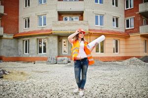 mujer ingeniera constructora con chaleco uniforme y casco protector naranja sostiene papel comercial contra el nuevo edificio. tema de bloque de vivienda de propiedad. foto