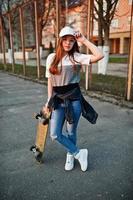 joven adolescente urbana con monopatín, gafas, gorra y jeans rotos en el campo de deportes del patio. foto