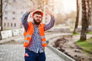 retrato de trabajador de barba brutal traje de hombre trabajador de la construcción en casco naranja de seguridad contra el pavimento mostrando los brazos.