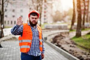 Portrait of beard worker man suit construction worker in safety orange helmet against pavement shows stop hand. photo