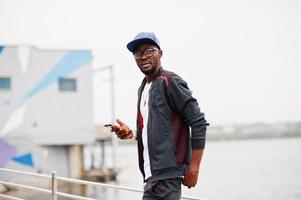 Portrait of stylish african american man on sportswear, cap and glasses with smartphone at hand. Black men model walk street against  lake. photo