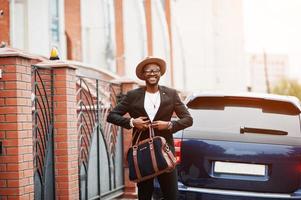 hombre negro con estilo en gafas con sombrero, vestido con traje con bolso contra coche de lujo. rico hombre de negocios afroamericano. foto