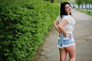 Back of sexy brunette girl on women's jeans shorts and white blouse against green spring bushes. photo