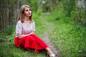 Portrait of sitting beautiful girl with red lips at spring blossom garden on green grass, wear on red dress and white blouse. photo