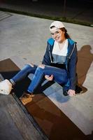 Young teenage urban girl with skateboard, wear on glasses, cap and ripped jeans at skate park on the evening. photo