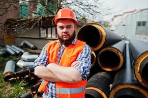 Brutal beard worker man suit construction worker in safety orange helmet near steel pipes. photo