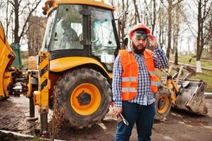 Brutal beard worker man suit construction worker in safety orange helmet, sunglasses against traktor with adjustable wrench at hand. photo