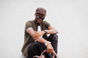 retrato de un elegante hombre afroamericano sentado con gafas de sol y gorra con bolso al aire libre. hombre negro de moda callejera. foto