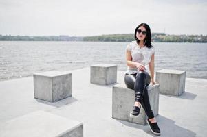 retrato de una chica morena escuchando música en los auriculares desde el teléfono móvil, usando pantalones de cuero de mujer y blusa blanca, gafas de sol, contra cubos de piedra en la playa del lago. foto