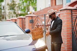 hombre negro con estilo en gafas con sombrero, vestido con traje con bolso contra coche de lujo. rico hombre de negocios afroamericano. mira los relojes. foto