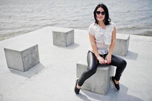 Portrait of brunette girl listening music at headphones from mobile phone, wear on women's leather pants and white blouse, sunglasses, against stone cubes at beach of lake. photo