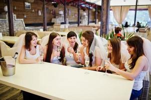 grupo de chicas alegres con camisas blancas sentadas en la mesa y bebiendo champán en la despedida de soltera.