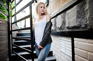 Elegante mujer rubia vestida con jeans, gargantilla y chaqueta de cuero en la calle cerca de las escaleras. retrato de modelo urbano de moda. foto